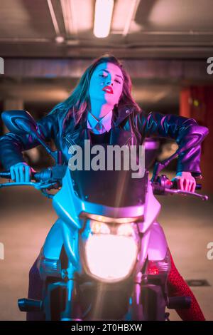 Fotografie mit blauen und rosa Neonfarben auf einem Motorrad. Porträt eines jungen blonden kaukasischen Models mit schwarzer Lederjacke Stockfoto