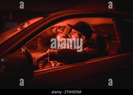 Mann in einer Kappe, der Auto fährt, raucht nachts in einer Garage, die mit rotem Licht beleuchtet ist, Sportwagen Stockfoto