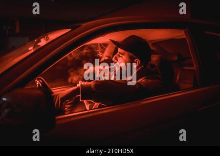 Mann in einer Kappe, der Auto fährt, raucht nachts in einer Garage, die mit rotem Licht beleuchtet ist Stockfoto