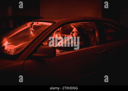 Rapper Mann in einer Kappe in einem Auto rauchen nachts in einer Garage beleuchtet mit rotem Licht im Winter, Sportwagen Stockfoto