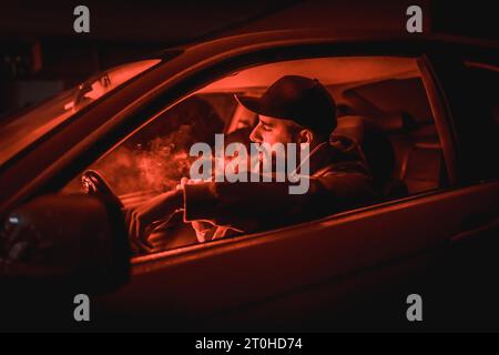 Mann in einer Kappe, der Auto fährt, raucht nachts in einer Garage, die mit rotem Licht beleuchtet ist Stockfoto