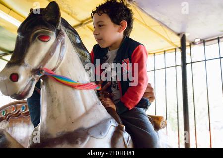 Venezolanisches Latino-männliches Kind, glücklich, mit seinem Vater auf einem vorgetäuschten Pferd auf der Karussellmesse zu reiten, Familienkonzept, Kopierraum. Stockfoto