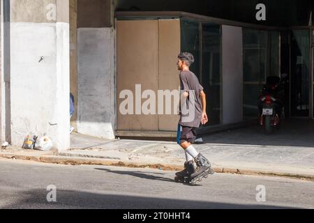 Ein junger Kubaner reitet Schlittschuhe auf einer Stadtstraße, wo ein kaputtes Schaufenster mit Brettern bedeckt ist. Auf dem Bürgersteig befinden sich Abschleppsäcke mit Müll. Stockfoto