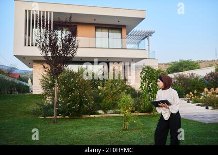 Eine Frau in einem Hijab nutzt ein intelligentes Tablet, um eine Villa im Hintergrund für den Verkauf vorzubereiten und dabei moderne Technologien bei der Vorbereitung von Immobilienlisten zu präsentieren Stockfoto