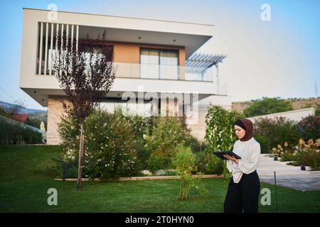 Eine Frau in einem Hijab nutzt ein intelligentes Tablet, um eine Villa im Hintergrund für den Verkauf vorzubereiten und dabei moderne Technologien bei der Vorbereitung von Immobilienlisten zu präsentieren Stockfoto