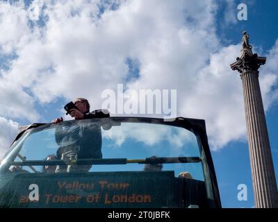 Gelber Londoner Tourbus vorbei an der Nelsons-Säule mit Touristen, die auf dem Oberdeck des offenen Busses vor blauem Himmel stehen und Fotos mit dem Telefon machen Stockfoto