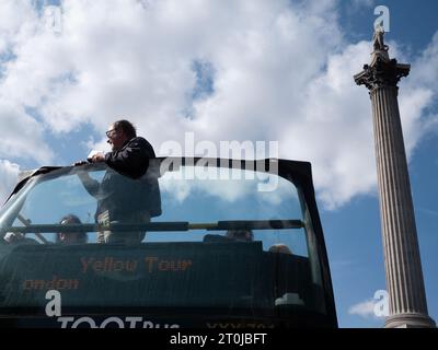 Gelber Londoner Tourbus vorbei an der Nelsons-Säule mit Touristen, die auf dem Oberdeck des offenen Busses vor blauem Himmel stehen Stockfoto
