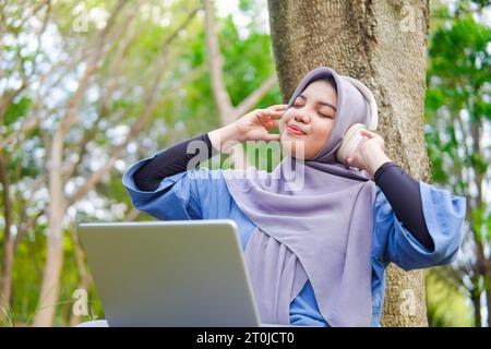 muslimische Frau mit Laptop und Kopfhörer. Hören Sie Musik im Park. Unter dem Strukturbaum Stockfoto