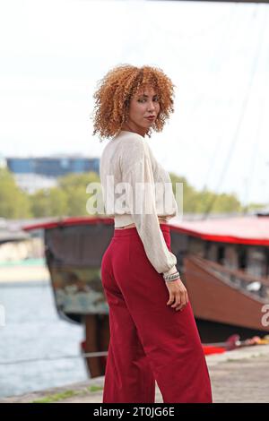 Frau mit lockigen Haaren und roten Hosen posiert auf einem Dock in der Nähe eines Bootes Stockfoto