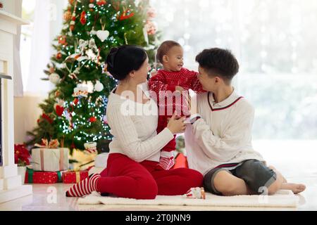 Asiatische Familie mit Kindern am Weihnachtsbaum und Kamin. Mutter und Kinder öffnen Geschenke am Kamin. Baby und Mutter öffnen Geschenke. Winterferien Stockfoto