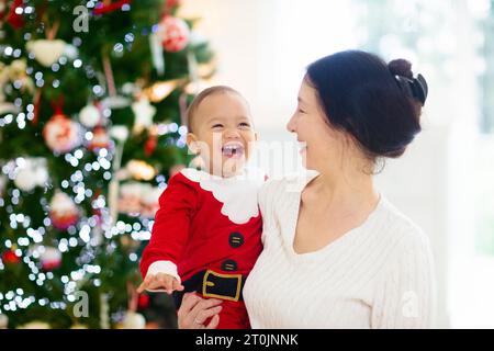 Asiatische Familie mit Kindern am Weihnachtsbaum und Kamin. Mutter und Kinder öffnen Geschenke am Kamin. Baby und Mutter öffnen Geschenke. Winterferien Stockfoto