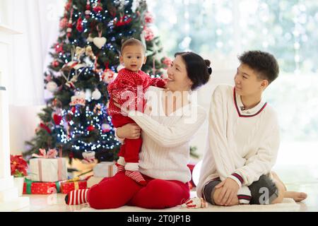 Asiatische Familie mit Kindern am Weihnachtsbaum und Kamin. Mutter und Kinder öffnen Geschenke am Kamin. Baby und Mutter öffnen Geschenke. Winterferien Stockfoto