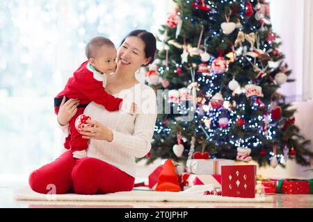 Asiatische Familie mit Kindern am Weihnachtsbaum und Kamin. Mutter und Kinder öffnen Geschenke am Kamin. Baby und Mutter öffnen Geschenke. Stockfoto