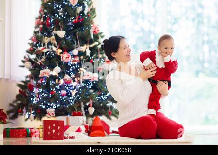 Asiatische Familie mit Kindern am Weihnachtsbaum und Kamin. Mutter und Kinder öffnen Geschenke am Kamin. Baby und Mutter öffnen Geschenke. Stockfoto