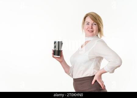 Reife blonde Frau, die einen schwarzen Einweg-Pappbecher für Kaffee oder Tee in der einen Hand hält, die andere Hand liegt auf der Taille. Frau, die in die Kamera schaut. Lächelnde Frau Stockfoto