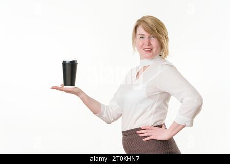 Lächelnde blonde Frau, die in der ausgestreckten Hand einen schwarzen Papierbecher für Kaffee hält und in die Kamera blickt. Zufriedene Frau 49 Jahre alt Stockfoto