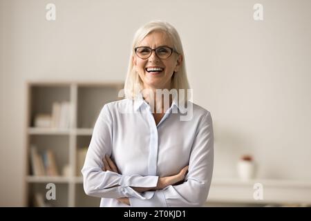 Fröhliche, selbstbewusste ältere Geschäftsfrau in eleganter Brille, die drinnen posiert Stockfoto