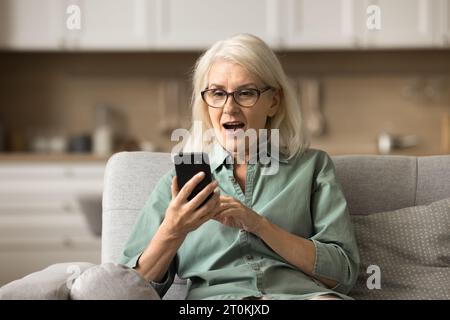 Überraschte alte Frau im Ruhestand mit Brille, die auf das Smartphone starrt Stockfoto