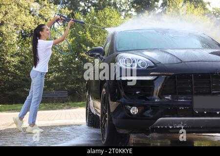 Glückliche Frau, die Auto mit Hochdruck-Wasserstrahl im Freien wäscht Stockfoto
