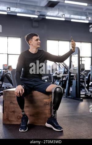 Sportler, der Videoanrufe macht. Der Kerl sitzt auf Holzkiste, macht Selfie im Fitnessstudio, Foto in voller Länge. Behinderter Mann, der im Sportzentrum fotografiert. Liebe Stockfoto