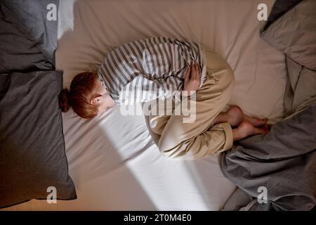 Über Draufsicht vertikales Bild in voller Länge stressige depressive, nervöse Ingwerfrau, die in fetaler Position auf dem Bett liegt, sich umarmt und an Schmerzen leidet Stockfoto