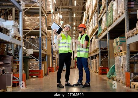 Junge kaukasische Arbeiter in Uniform machen eine Bestandsverwaltung von Produkten in Regalen im Lager, diskutieren gemeinsam. Konzept eines guten Managementsystems Stockfoto