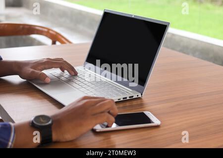 Nahaufnahme eines Geschäftsmannes, der den leeren Bildschirm des Mobiltelefons berührt, mit einem Laptop neben dem Tisch. Display von Smartphone und Laptop-Modell Stockfoto