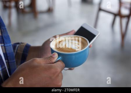 Nahaufnahme eines asiatischen Mannes, der eine Tasse Kaffee und ein Handy hält. Technik und morgendliches Routinekonzept Stockfoto