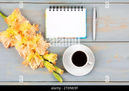 Künstlerische Komposition mit offenem leerem Notizblock und Stift, Tasse schwarzen Kaffee, orange-gelbe Gladiolusblüte auf blauem Holztisch. Feierlicher Bürotisch Stockfoto