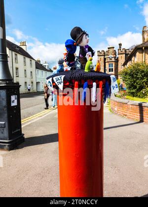Ein gestrickter Topper-Hut mit Figuren auf einem roten königlichen Briefkasten, der an einem sonnigen Tag eine skurrile Szene an einer Straßenecke macht. Stockfoto