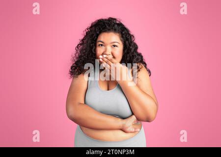 Oh, Junge schwarze Frau in Übergröße, die Sportkleidung trägt, posiert und den Mund mit Handfläche bedeckt, steht auf rosa Hintergrund Stockfoto