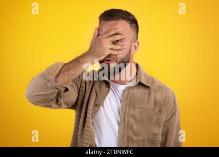 Ein lächelnder, kaukasischer Typ mit Bart in der Freizeit bedeckt Gesicht mit Hand, Spione mit Auge Stockfoto