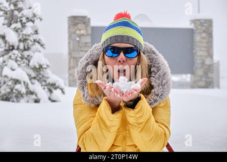 Frau, die Schnee aus den Händen bläst, im Winter farbenfrohe gelbe Kleidung trägt. Glück, Freude, verspielt. Stockfoto