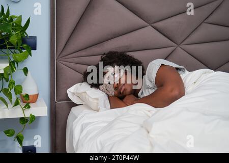Afrikanische junge Frau, die mit einer niedlichen Schlafmaske auf weißen Laken im Bett in einem Zimmer mit einer Pflanze schläft. Schlafen. Stockfoto