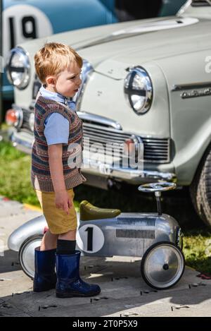 Ein kleiner Junge im Vintage-Kostüm mit einem klassischen großen Spielzeugauto und einem großen Oldtimer beim Goodwood Revival Nostalgie Event. Retro Stockfoto