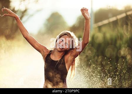 Im Hinterhof des Hauses platzt ein junges Mädchen vor Lachen und Freude, als sie an einem sonnigen Sommertag fröhlich durch einen Wassersprenger rennt Stockfoto