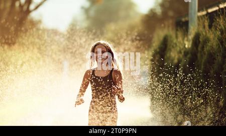 Im Hinterhof des Hauses platzt ein junges Mädchen vor Lachen und Freude, als sie an einem sonnigen Sommertag fröhlich durch einen Wassersprenger rennt Stockfoto