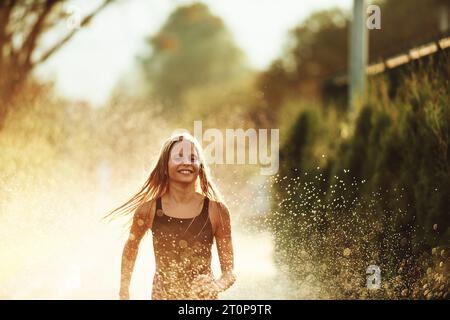 Im Hinterhof des Hauses platzt ein junges Mädchen vor Lachen und Freude, als sie an einem sonnigen Sommertag fröhlich durch einen Wassersprenger rennt Stockfoto