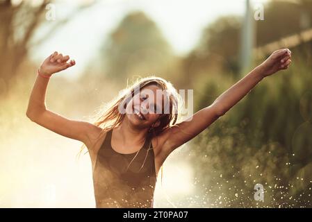 Im Hinterhof des Hauses platzt ein junges Mädchen vor Lachen und Freude, als sie an einem sonnigen Sommertag fröhlich durch einen Wassersprenger rennt Stockfoto