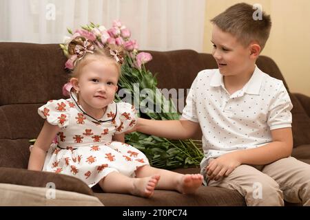 Familienkonzept. Bruder und Schwester spielen glücklich, während sie auf einem braunen Sofa sitzen, in einem Wohnraum vor dem Hintergrund eines rosa Blumenstraußes. Stockfoto