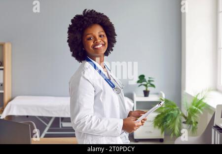 Glücklicher afroamerikanischer Arzt im Labormantel, der in der Arztpraxis steht Stockfoto