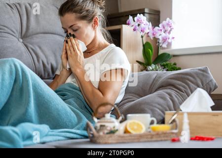 Frau niest in ein Taschentuch. Eine kranke Frau, die zu Hause unter einer Decke sitzt. Vor ihr steht ein Tablett mit einer Teekanne, Zitrone und Honig. Stockfoto