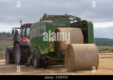 Eine McHale-Ballenpresse, die von einem Red Valtra-Traktor gezogen wird und während der Ernte von hinten gesehen zwei Strohballen auf einem Stoppelfeld platziert Stockfoto