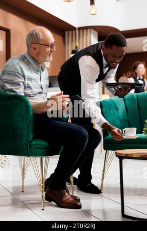 Im Loungebereich gab es einen netten Kellner, der älteren männlichen Touristen eine Tasse Tee servierte, die auf der Couch mit Tablet saßen. Seniorenmann im Ruhestand, der auf einem gemütlichen Sofa sitzt und von afroamerikanischen Mitarbeitern in der Hotellobby besucht wird. Stockfoto
