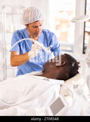 Afrikanischer Mann unterzieht sich einer Feuchtigkeitsbehandlung der Gesichtshaut mit Wasserstoff-Sauerstoff-Hydratationsdüse Stockfoto