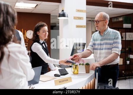 Ein pensionierter Senior-Mann meldet sich mit Hilfe eines Concierge an einer Hotelrezeption an. Älterer männlicher Tourist zahlt mit einer Kreditkarte an einem NFC-Terminal, während er sein Gepäck hält. Stockfoto