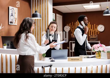 Die Rezeptionisten an der Rezeption helfen Senioren beim Check-in. Während das Personal Anrufe entgegennimmt und eine reibungslose Abwicklung ermöglicht, unterschreibt eine dankbare ältere Frau Papiere mit einem Lächeln. Ausgezeichneter Kundenservice. Stockfoto