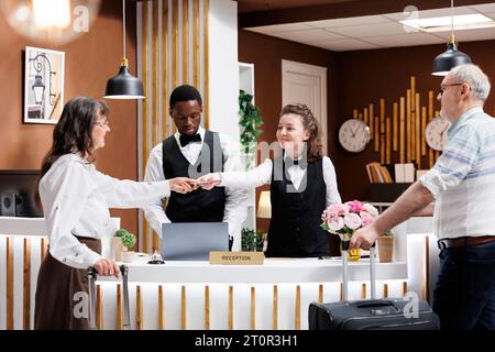 Der freundliche Concierge hilft älteren Touristen in der Hotellobby. Pensionierte Seniorin stellt die notwendigen Dokumente für den Check-in zur Verfügung, während ein alter Reisender geduldig an der Rezeption wartet. Stockfoto