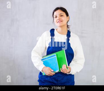 Junge Mechanikerin in blauen Overalls, die Arbeitsbücher hält Stockfoto