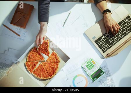 Nachhaltiger Arbeitsplatz. Obersicht einer Arbeitnehmerin im grünen Büro mit Pizza und Laptop. Stockfoto